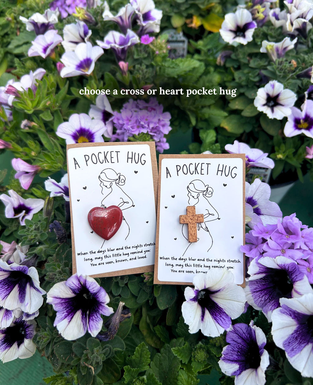 Two small 'A Pocket Hug' for a new mom carded gift items displayed among purple and white flowers. One features a red heart-shaped pocket, and the other features a wooden cross-shaped pocket.