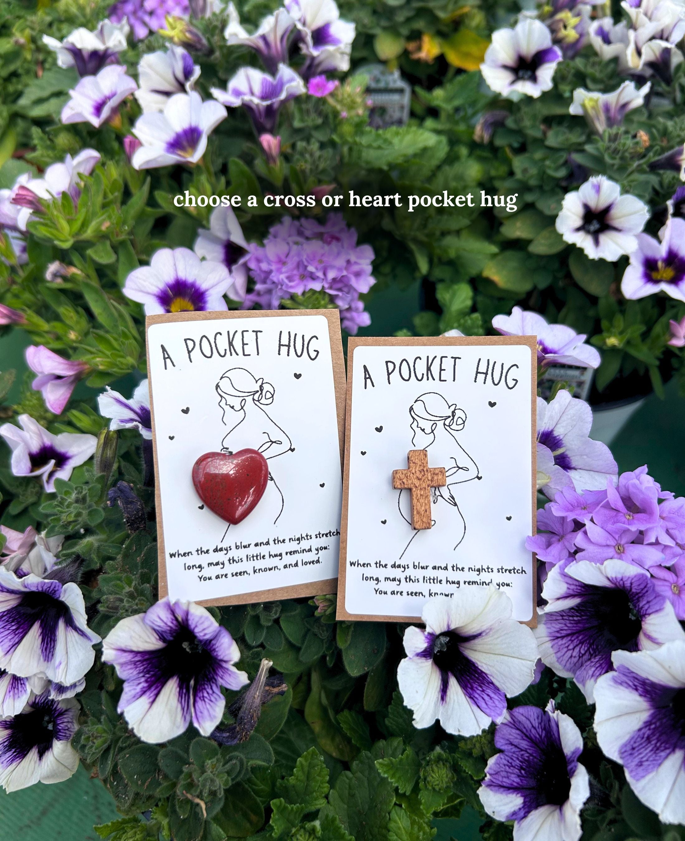 Two small 'A Pocket Hug' for a new mom carded gift items displayed among purple and white flowers. One features a red heart-shaped pocket, and the other features a wooden cross-shaped pocket.