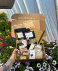 A hand holds an open cardboard postpartum gift box filled with assorted tea products, small packaged items, and handwritten notes, set against a garden background of flowers and greenery.