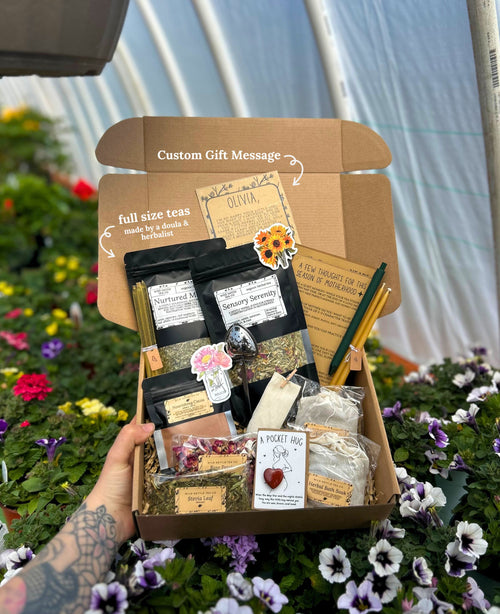 A hand holds an open cardboard postpartum gift box filled with assorted tea products, small packaged items, and handwritten notes, set against a garden background of flowers and greenery.
