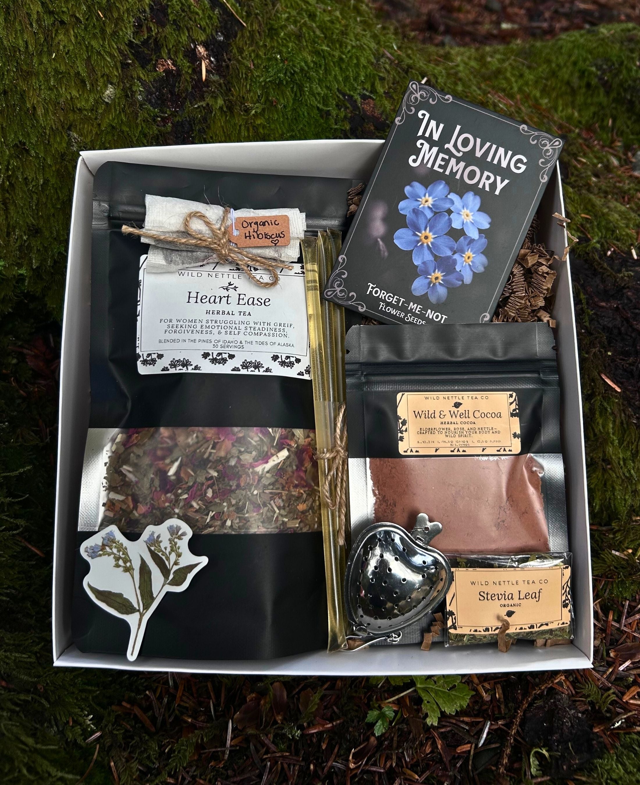 A curated sympathy gift for loss. A tea gift box set displayed in a white tray on mossy ground. The set includes a black card reading 'In Loving Memory', 'Heart Ease' herbal tea, 'Wild Ginger Co' tea, 'Stesia Leaf' tea, a silver heart-shaped tea infuser, dried flowers, and a white floral sticker.