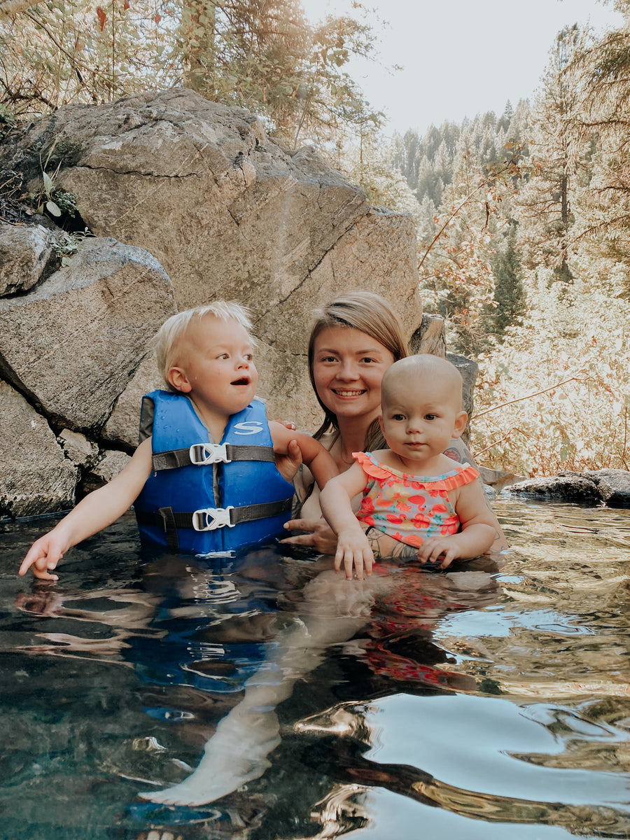 Hanna smiling in a forest pool, holding her baby in a coral swimsuit while her toddler in a blue life vest points at the water, with boulders and pine trees behind them.