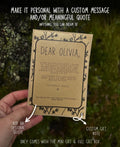 A hand holds a personalized mini gift card with a quote for 'Olivia' against a blurred green outdoor background. White text overlays explain that the card can be personalized with a custom message and quote, and point out features like adding an optional quote and a custom gift note.