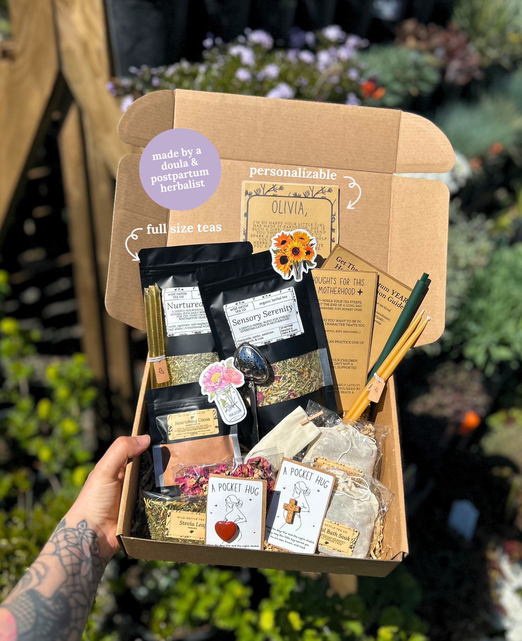 A hand holds an open kraft gift box filled with tea products for a new mom gift basket, sachets, cards, and accessories outdoors. The box includes black tea bags, loose herbal blends, a metal spoon, wooden tools, and personalized labels.