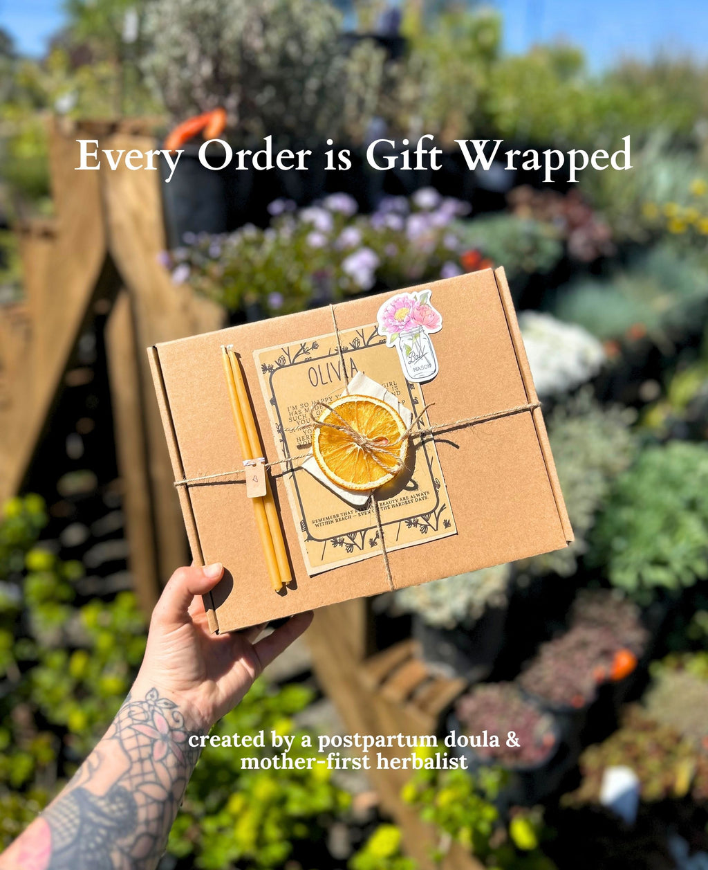 A hand holds a brown kraft gift box wrapped with twine and decorated with a dried orange slice, a small yellow candle, and a floral sticker, set against a blurred garden background. Text overlays read 'Every Order is Gift Wrapped' and 'created by a postpartum doula & mother-first herbalist'.