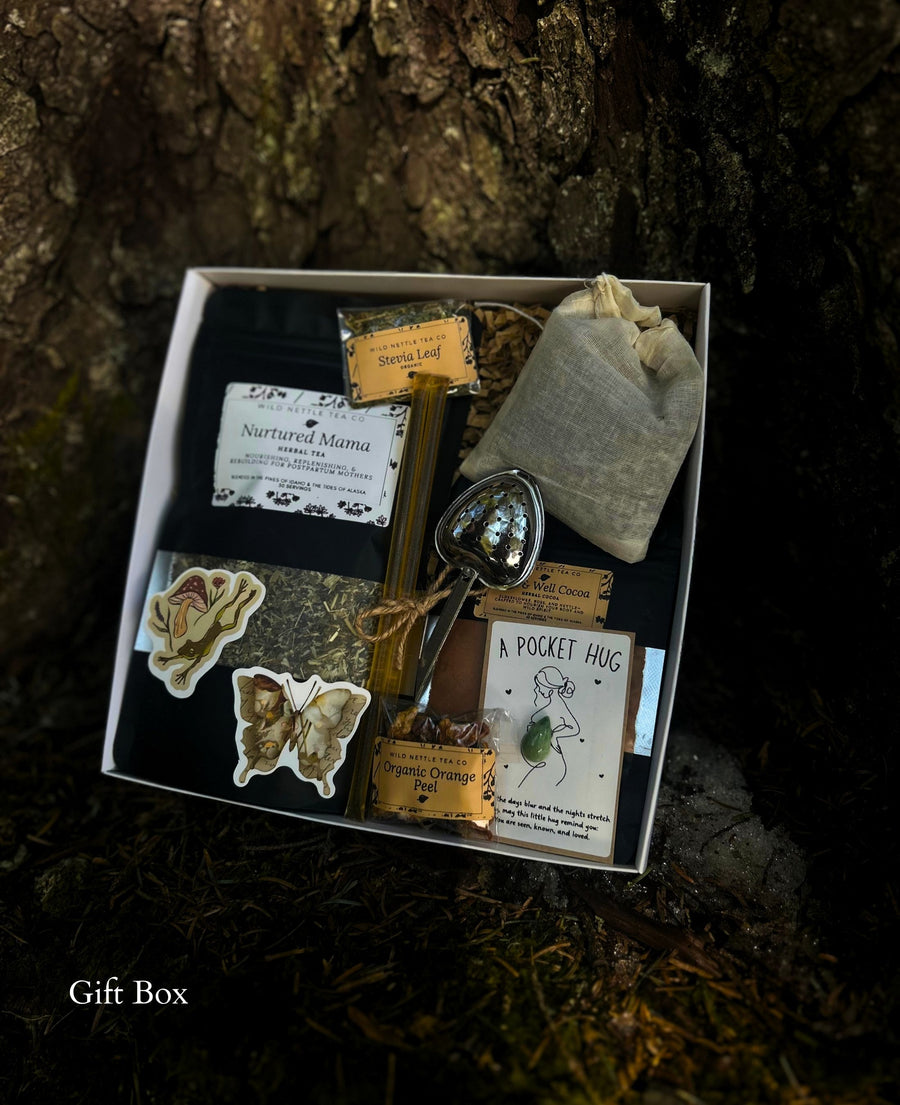 A curated nature-themed postpartum gift basket set displayed on a dark forest floor in front of a tree trunk. The white box contains a silver metal tea infuser, a small beige fabric pouch, dried botanicals, a tea bag, a butterfly-shaped sticker, a flower sticker, and a card labeled 'A POCKET HUG'.