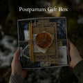 A person holds a postpartum gift box with wrapping in an outdoor setting. The box contains a dried orange slice, a candle, a small bottle, and decorative paper elements tied together with twine.
