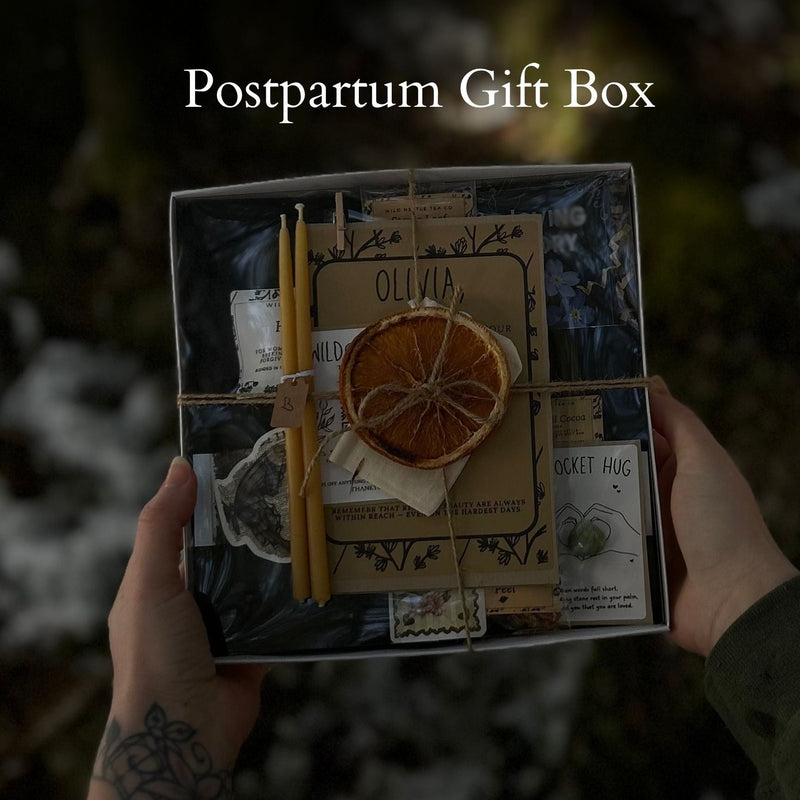 A person holds a postpartum gift box with wrapping in an outdoor setting. The box contains a dried orange slice, a candle, a small bottle, and decorative paper elements tied together with twine.