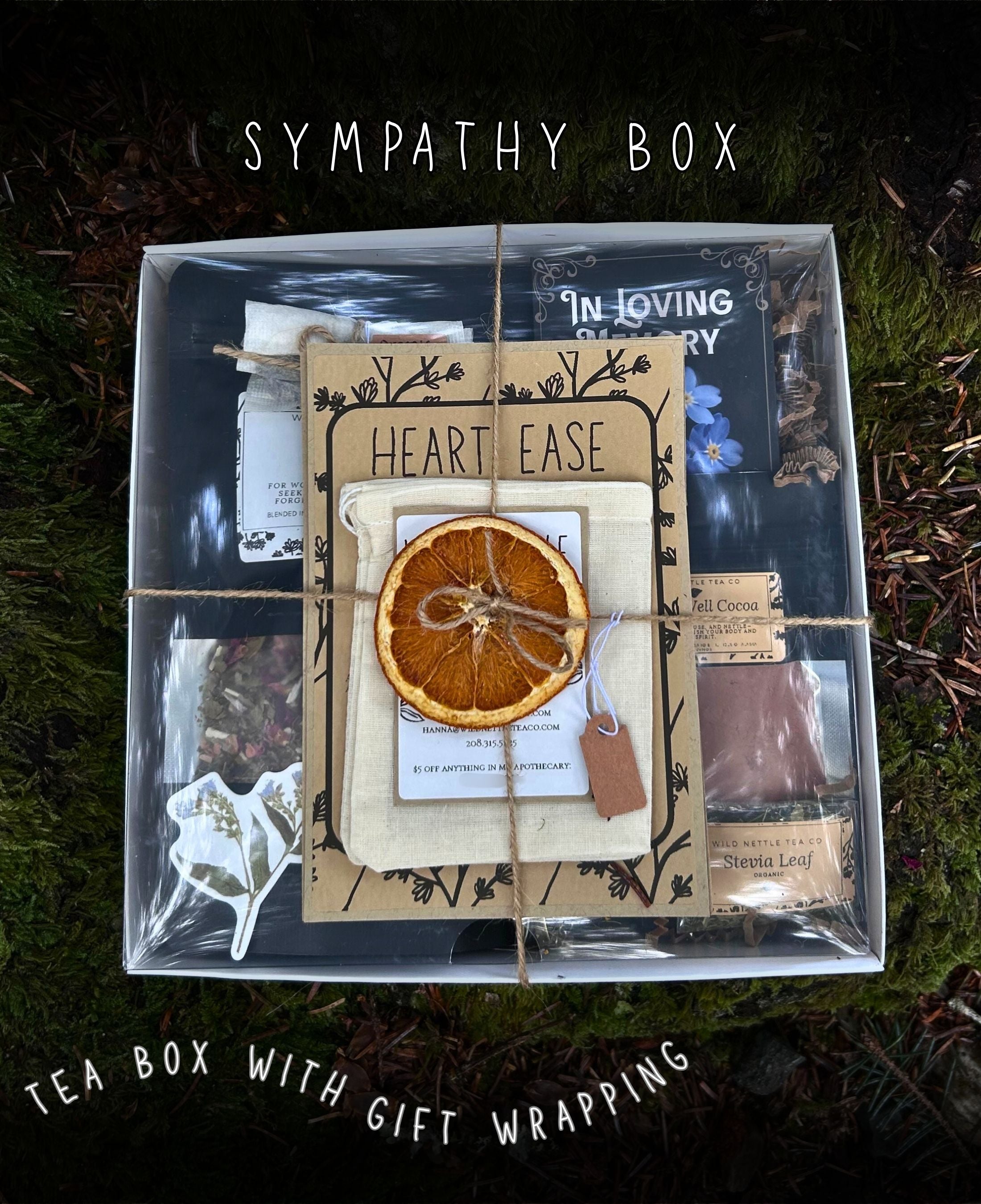 A clear plastic gift box containing assorted tea items and wrapping materials, placed outdoors on mossy ground. The box is wrapped with twine and layered with cards, a dried orange slice, and small tea bags.