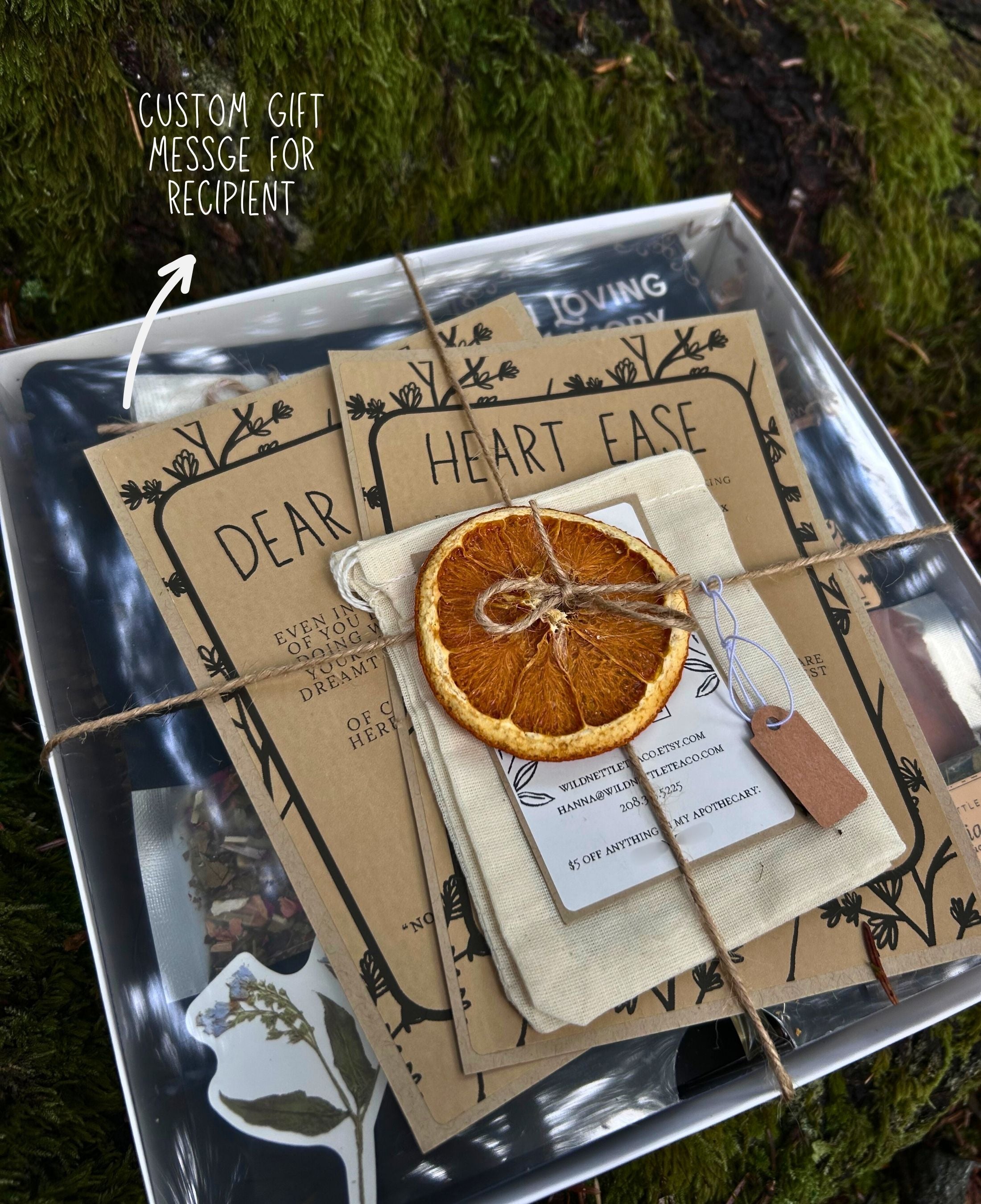 A sympathy gift box arrangement featuring kraft paper cards with black botanical-style borders, a small cream fabric pouch tied with twine, a dried orange slice, and a small tag. The items are placed on a reflective silver tray outdoors on mossy ground, with a handwritten note in the upper left reading 'Custom Gift Message for Recipient'.