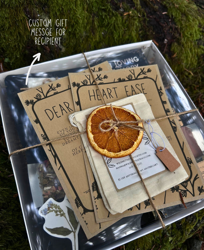 A sympathy gift box arrangement featuring kraft paper cards with black botanical-style borders, a small cream fabric pouch tied with twine, a dried orange slice, and a small tag. The items are placed on a reflective silver tray outdoors on mossy ground, with a handwritten note in the upper left reading 'Custom Gift Message for Recipient'.