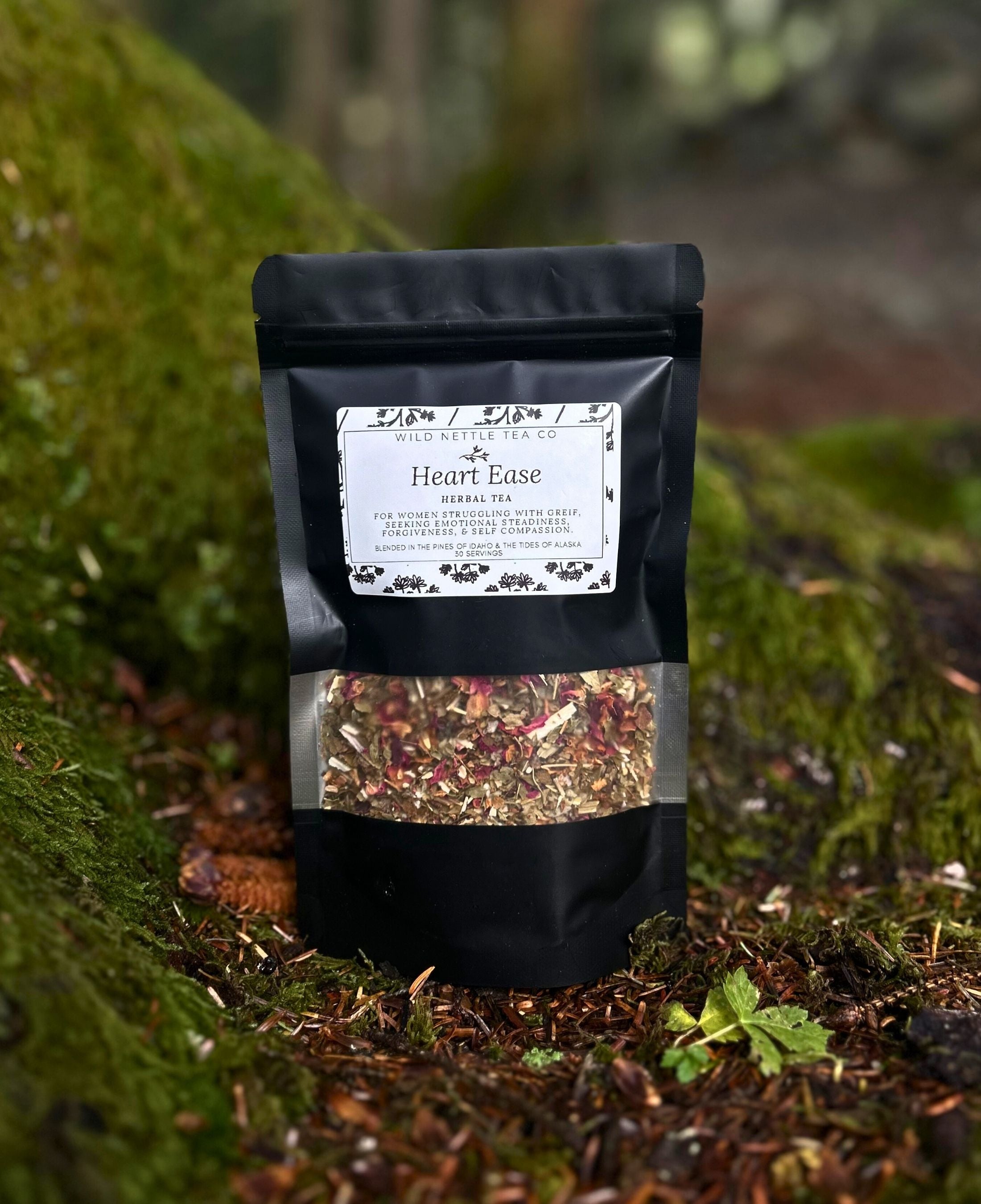 A black stand-up pouch of Heart Ease herbal tea from Your Friend Hanna, placed on mossy forest ground with a blurred natural background.