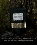 A black stand-up pouch labeled 'Whole Mother' organic herbal tea, leaning against a moss-covered tree trunk outdoors. The pouch has a clear window showing a loose leaf herbal blend and a white label with black text.
