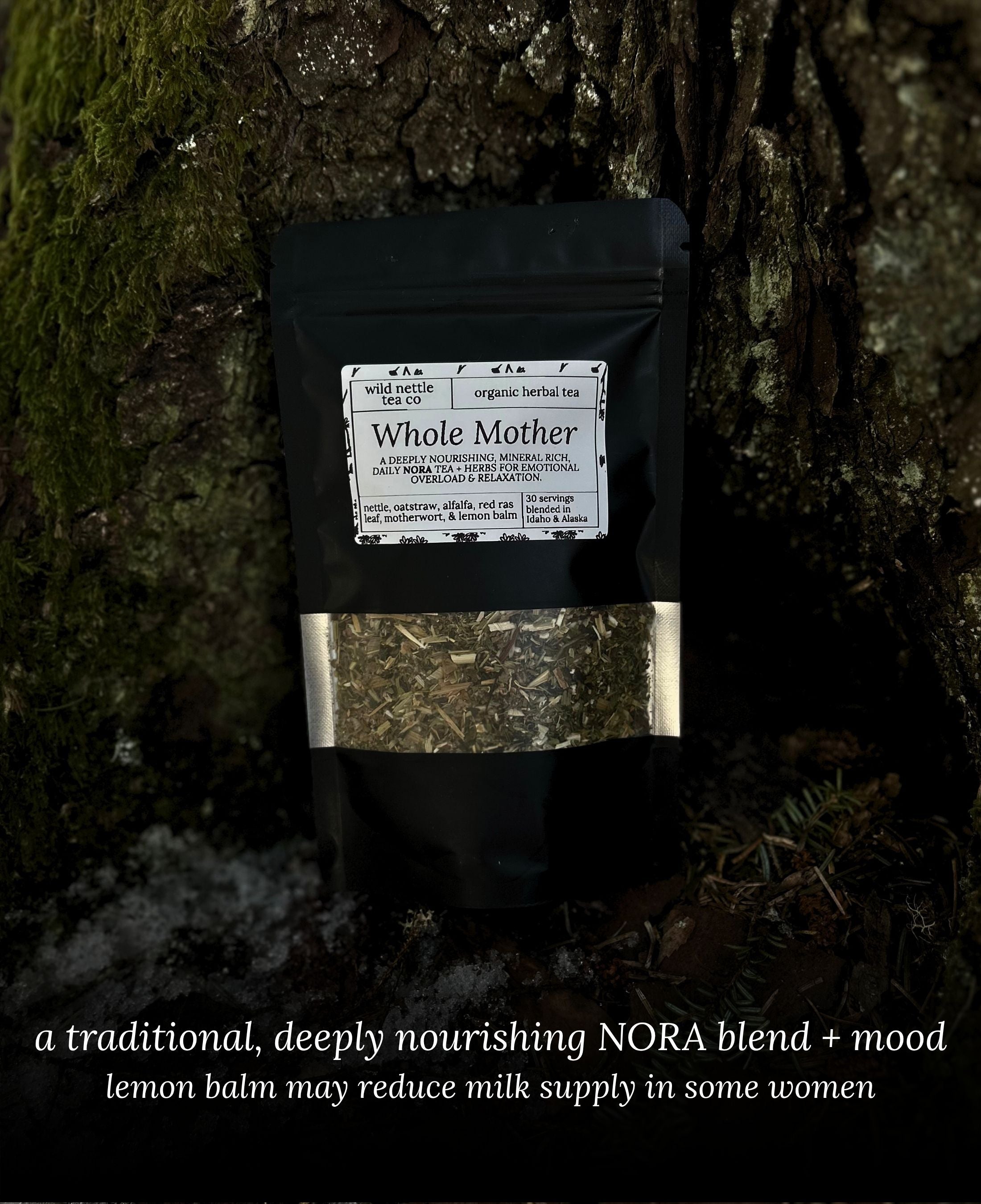 A black stand-up pouch labeled 'Whole Mother' organic herbal tea, leaning against a moss-covered tree trunk outdoors. The pouch has a clear window showing a loose leaf herbal blend and a white label with black text.